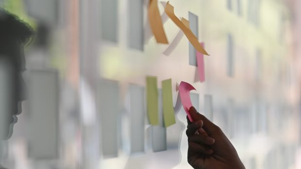 Side shot of a technology professional putting a sticky note on a reflecitive glass wall. The concept of self-reflection coaching techniques is implied.